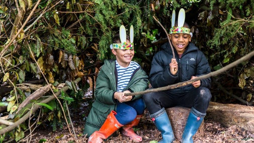 Two boys sit in a woodland den wearing bunny ears